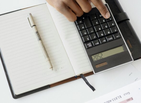 A hand using a calculator with financial documents and notebook, depicting an office setup for financial analysis.